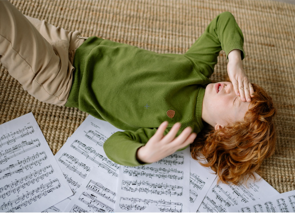 01 Person with red hair wearing a green sweater lies on the floor surrounded by sheet music resting their head on their hands.